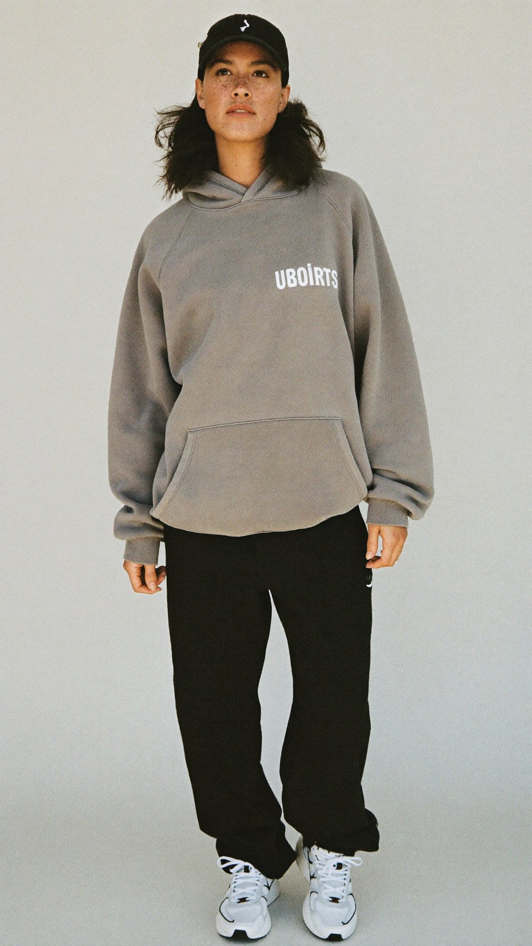 Full-body reference photo of the AI character: a woman in a black cap and grey hoodie